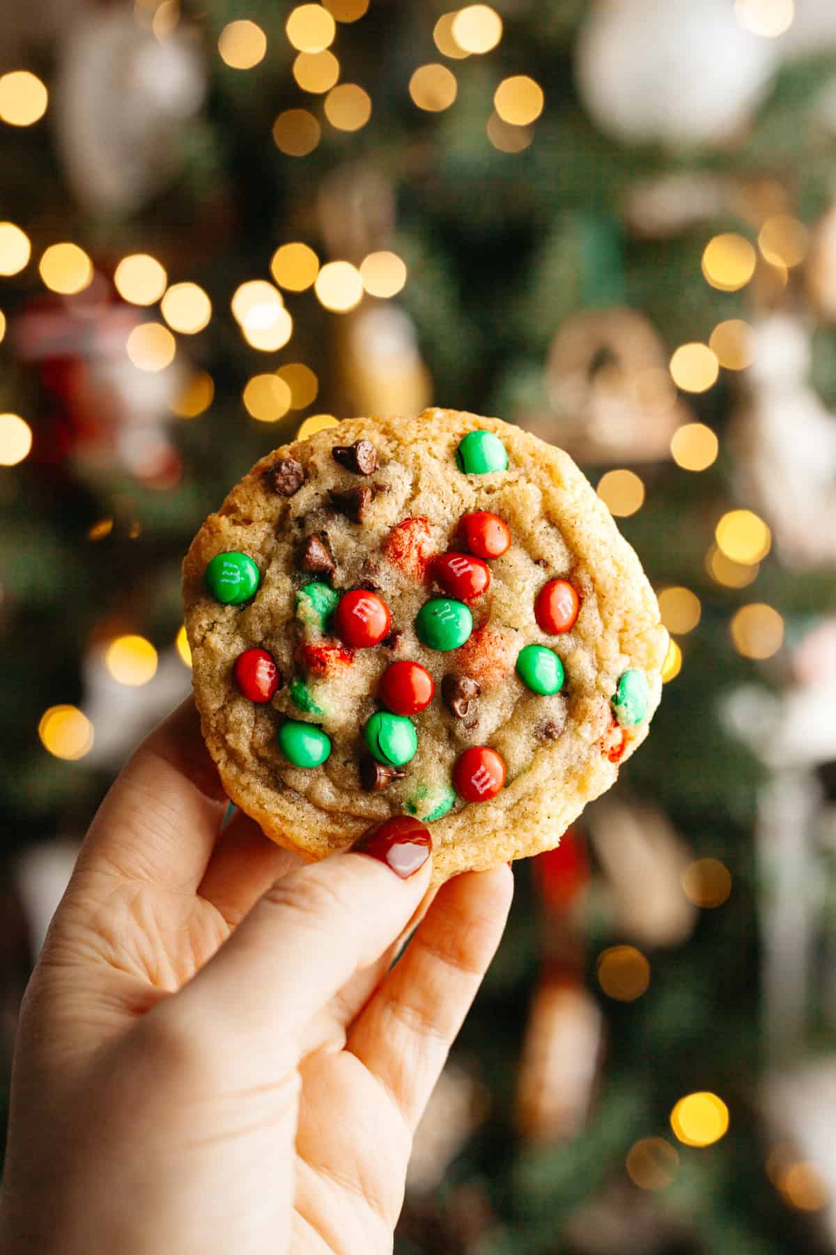 A hand holding a red and green M&M chocolate chip cookie.