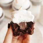 A hand holding a chocolate cupcake with a bite taken out of it, it has white frosting and a chocolate ganache filling.