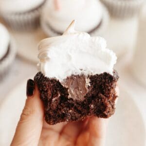 A hand holding a chocolate cupcake with a bite taken out of it, it has white frosting and a chocolate ganache filling.