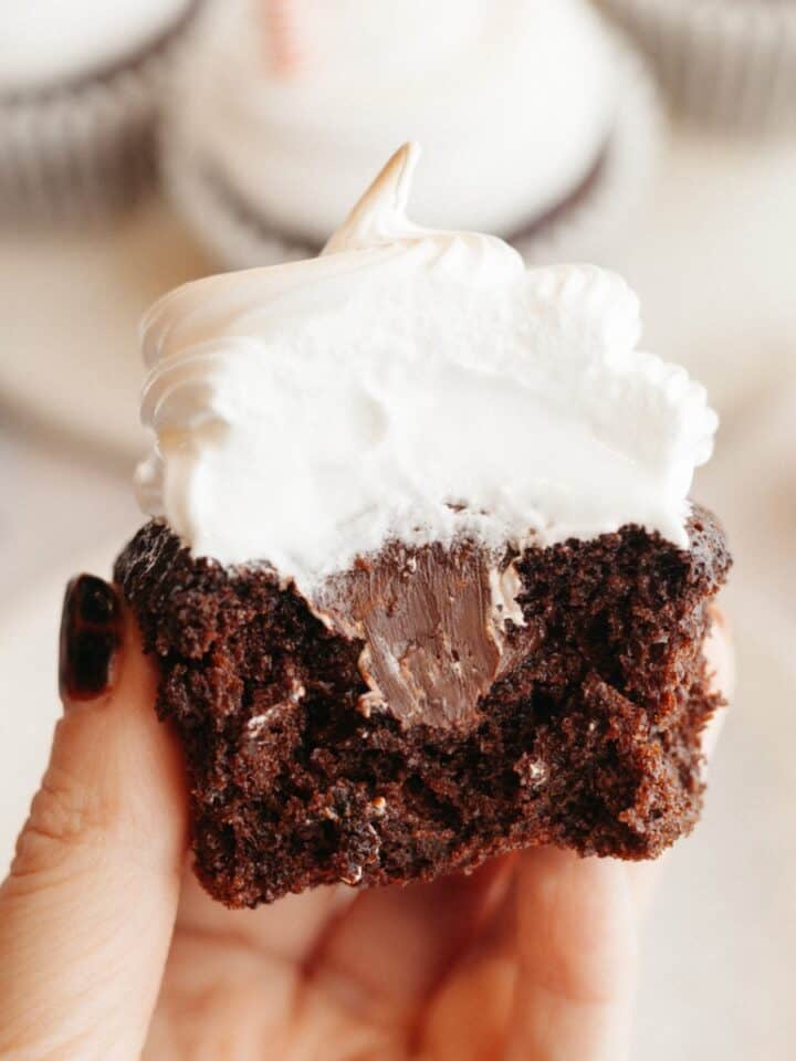 A hand holding a chocolate cupcake with a bite taken out of it, it has white frosting and a chocolate ganache filling.