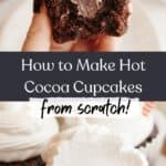 A hand holding a chocolate cupcake with a bite taken out of it, it has white frosting and a chocolate ganache filling, the same cupcake is on a plate.