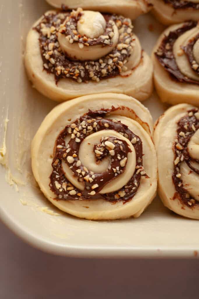 A close up of unbaked Nutella cinnamon rolls in a white dish.