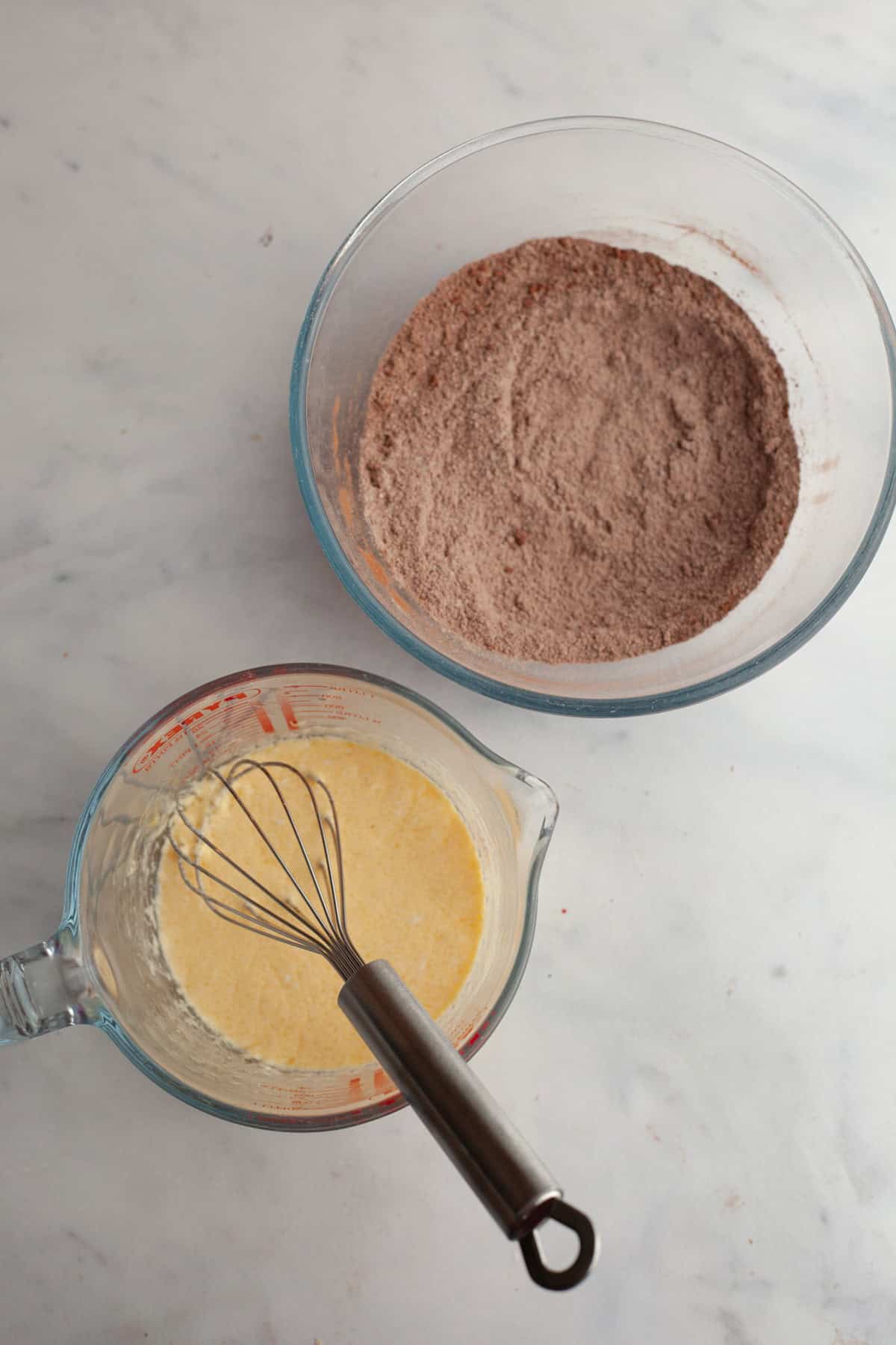 A glass bowl with flour and cocoa powder and a small jug with whisked eggs in it.