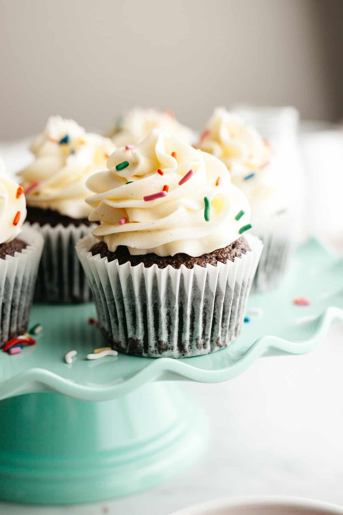 Chocolate cupcakes with vanilla frosting and rainbow sprinkles on a teal cake stand.