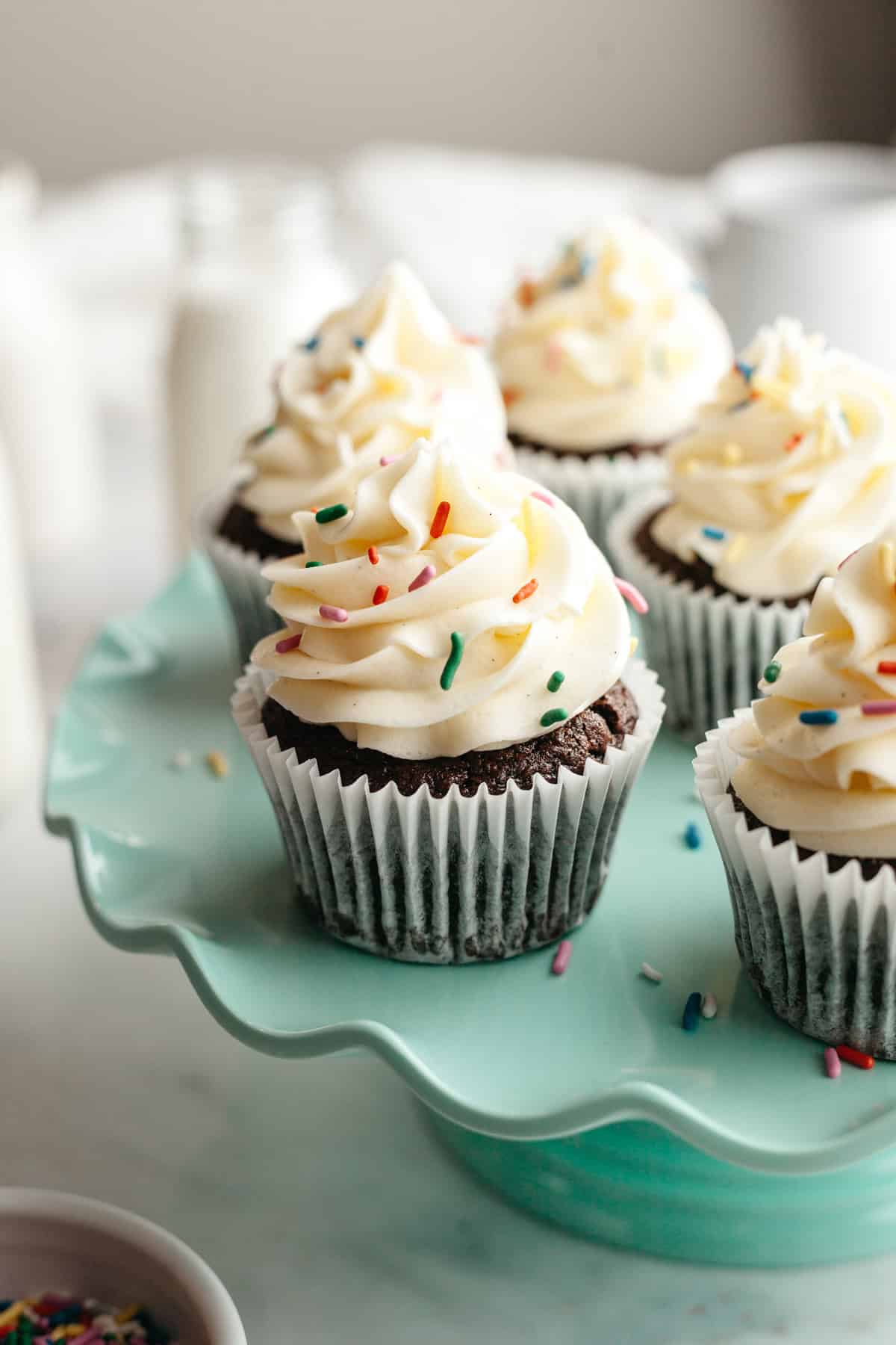 Chocolate cupcakes with vanilla frosting and rainbow sprinkles on a teal cake stand.