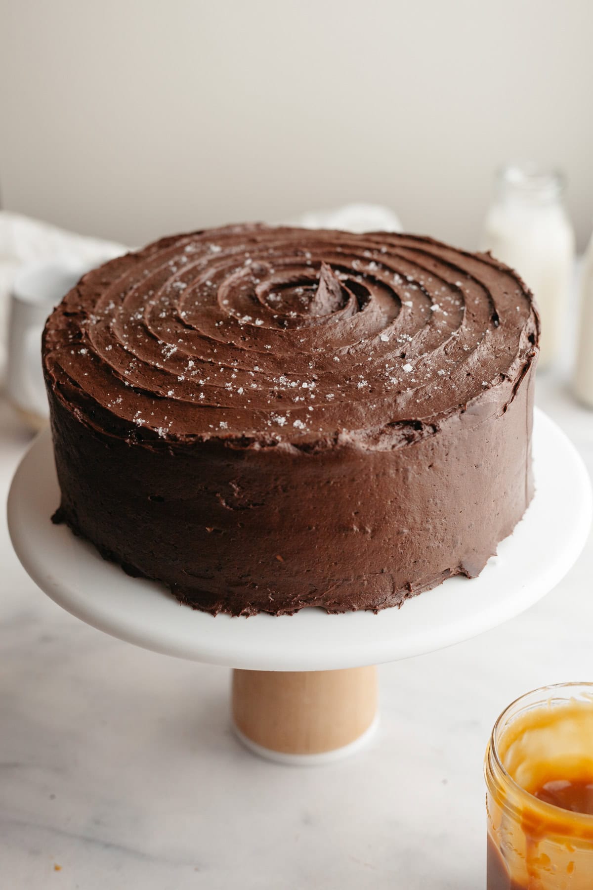 A white cake stand with a chocolate frosted cake on top.