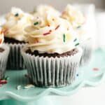 Chocolate cupcakes with vanilla frosting and rainbow sprinkles on a teal cake stand.