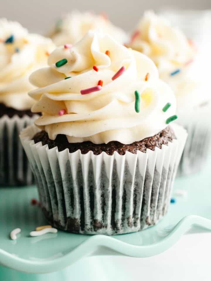 Chocolate cupcakes with vanilla frosting and rainbow sprinkles on a teal cake stand.