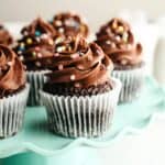 Chocolate cupcakes with chocolate frosting and rainbow sprinkles on a teal cake stand.
