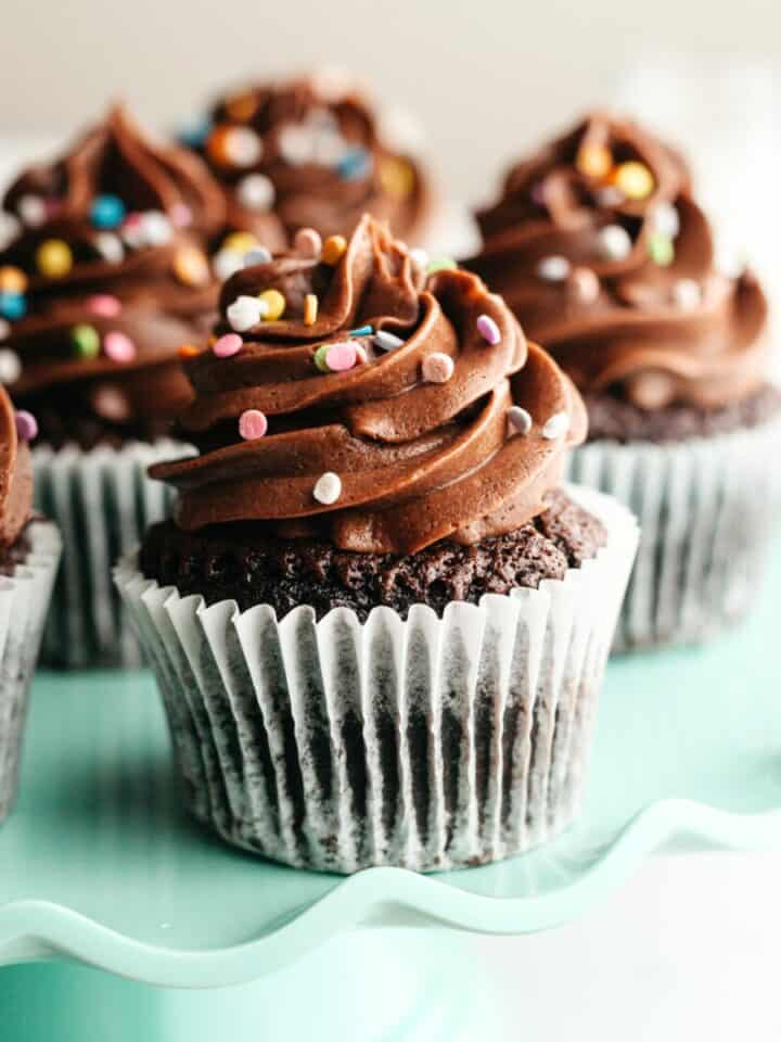 Chocolate cupcakes with chocolate frosting and rainbow sprinkles on a teal cake stand.