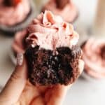 A hand holding a chocolate cupcake with strawberry buttercream with a bite taken out of it, showing a jam middle.