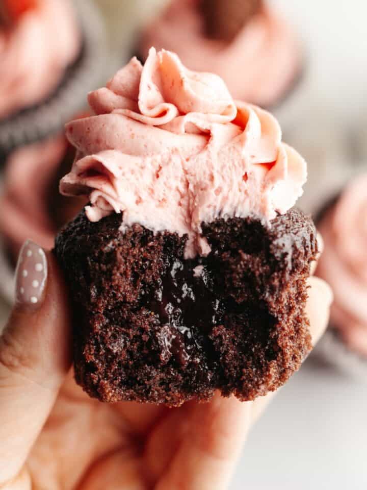 A hand holding a chocolate cupcake with strawberry buttercream with a bite taken out of it, showing a jam middle.