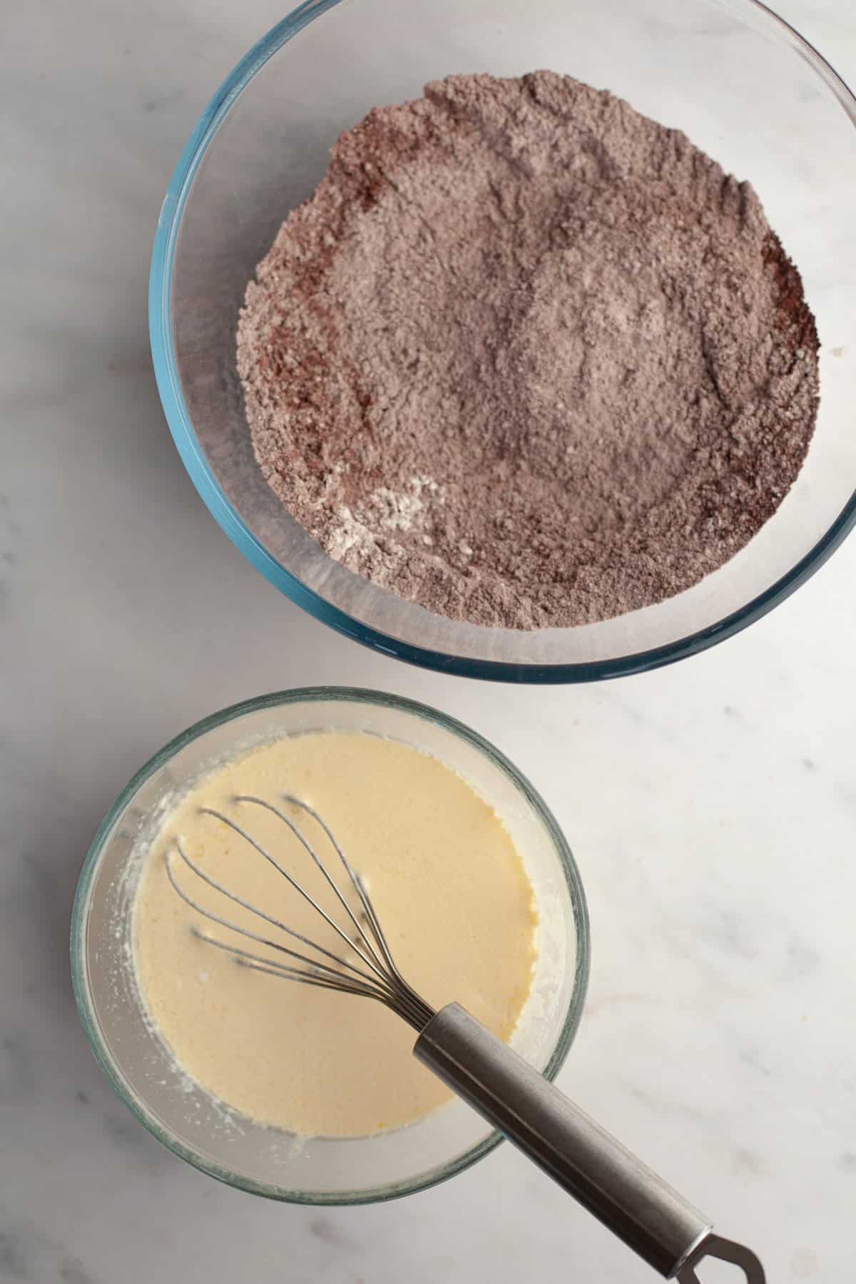 A large bowl with flour and cocoa powder, and a small bowl with a whisk and a pale yellow mixture.