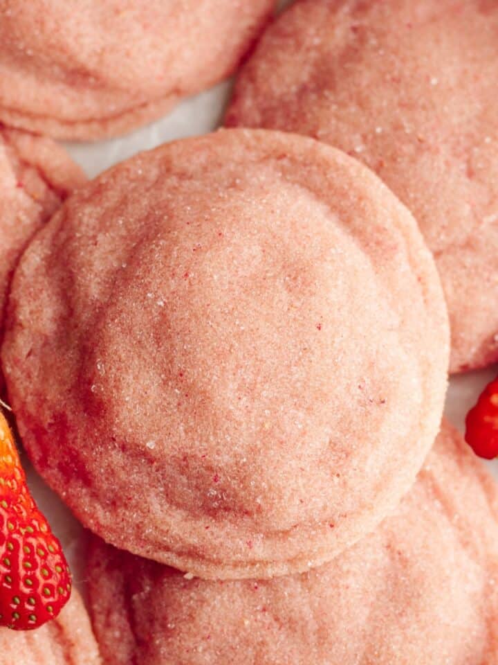 A close up of pink strawberry sugar cookies on parchment paper, there are a few sliced strawberries.