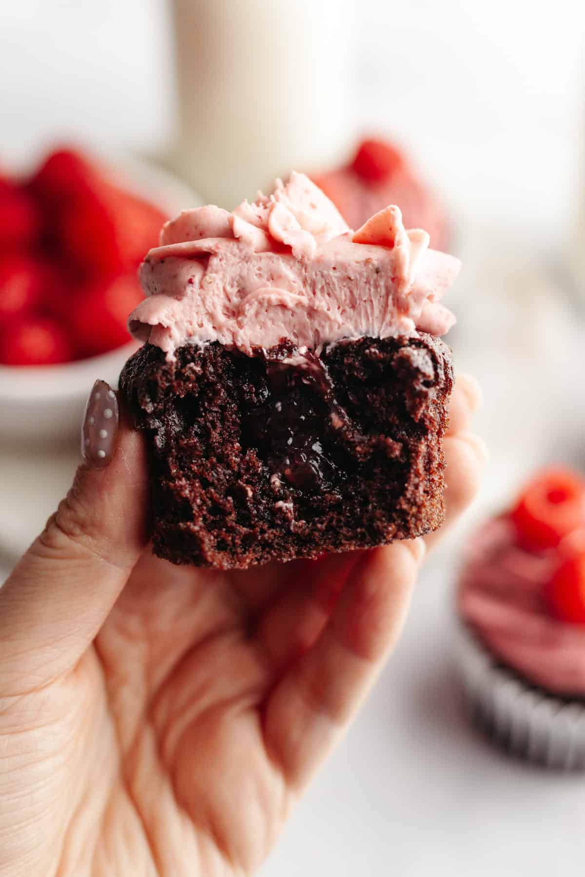 A hand holding a chocolate cupcake with raspberry frosting, a bite has been taken and shows a raspberry jam filling.