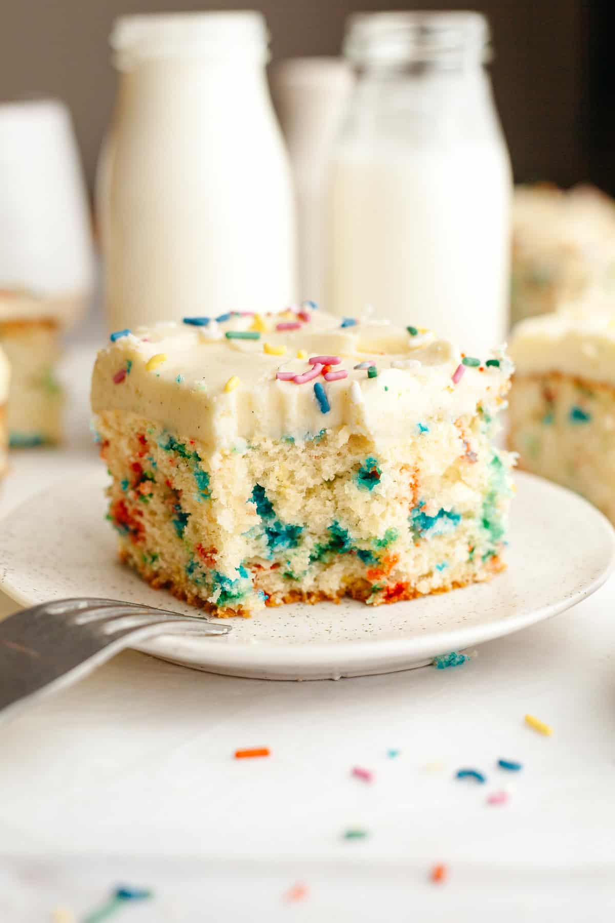 A slice of confetti sheet cake with vanilla frosting, a bite has been taken out of it.