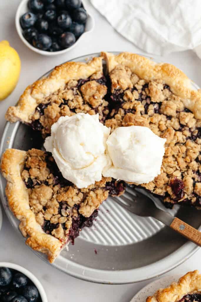 Blueberry crumble topped pie in a silver pie dish, topped with two scoops of vanilla ice cream.