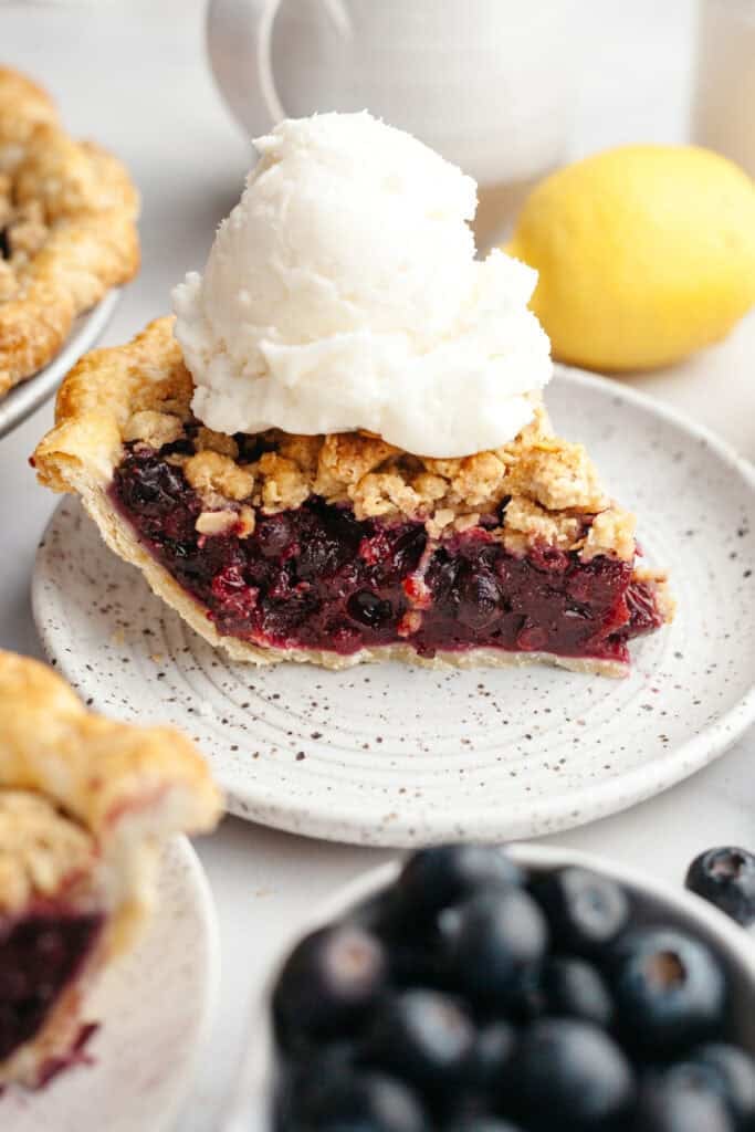 A slice of blueberry crumble top pie with a large scoop of vanilla ice cream.