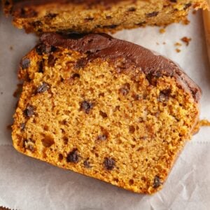A close-up of a slice of chocolate chip pumpkin loaf cake on white parchment paper, showing its moist texture and chocolate chips.