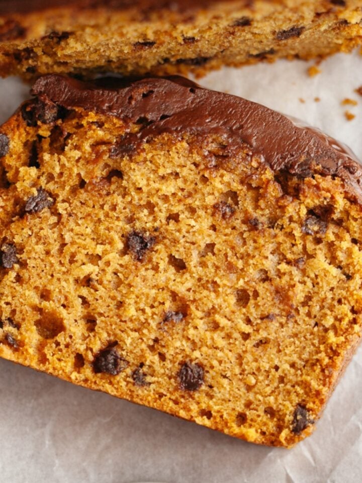 A close-up of a slice of chocolate chip pumpkin loaf cake on white parchment paper, showing its moist texture and chocolate chips.