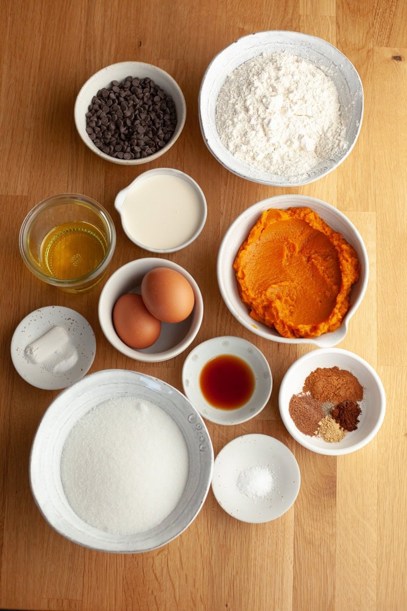 Ingredients on a wooden surface: flour, pumpkin puree, chocolate chips, eggs, milk, oil, sugar, vanilla, baking soda, spices, and salt, each in separate bowls, viewed from above.