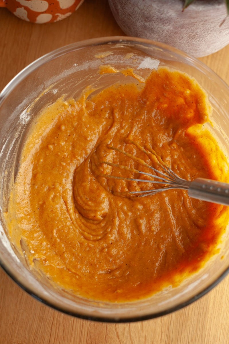 A wooden table with a glass bowl filled with pumpkin batter and a whisk.
