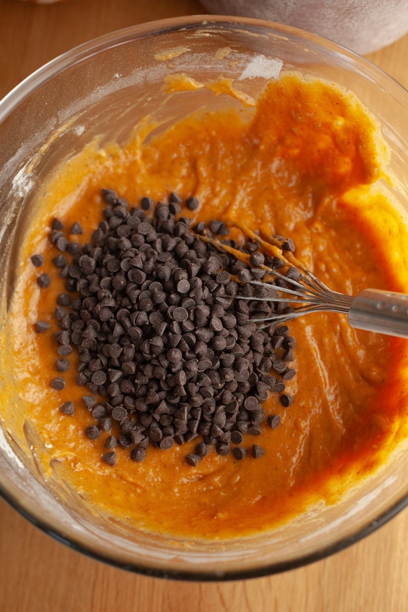A wooden table with a glass bowl, with pumpkin loaf batter and mini chocolate chips.