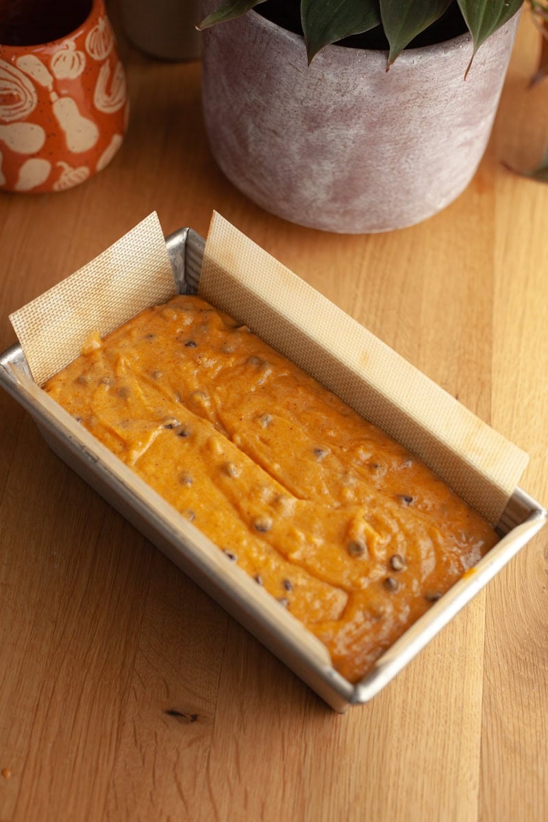 A loaf pan lined with parchment paper is filled with pumpkin bread batter that has chocolate chips mixed in, sitting on a wooden surface near potted plants.