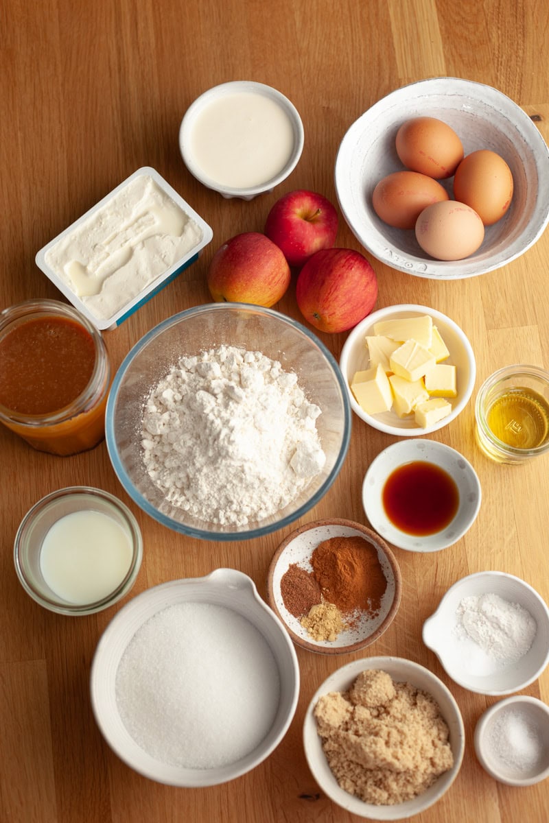 A top-down view of baking ingredients on a wooden surface, including flour, sugar, eggs, apples, butter, milk, vanilla, cinnamon, brown sugar, cream cheese, oil, and baking powder.
