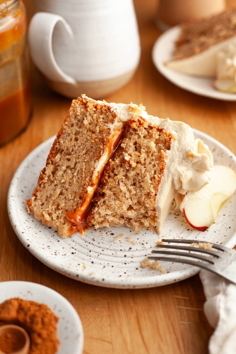 A close-up of a slice of apple layered cake with cream cheese frosting and apple garnish, served on a speckled plate. A bite has been taken out of it with a fork.