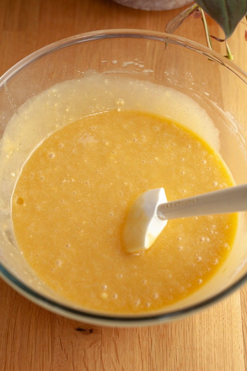 A glass bowl on a wooden table, the bowl is filled with yellow cake batter and a rubber spatula.