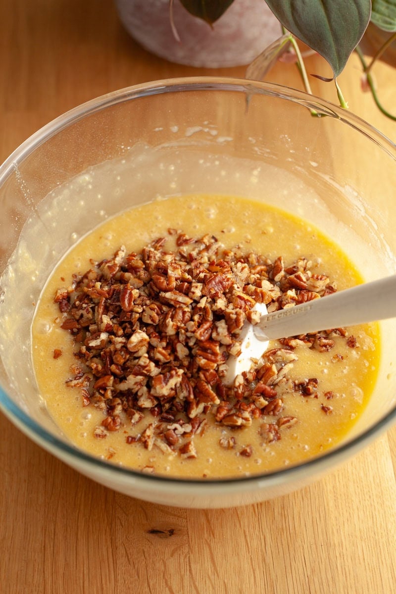 A glass bowl on a wooden table, the bowl is filled with yellow cake batter, chopped pecans, and a rubber spatula.