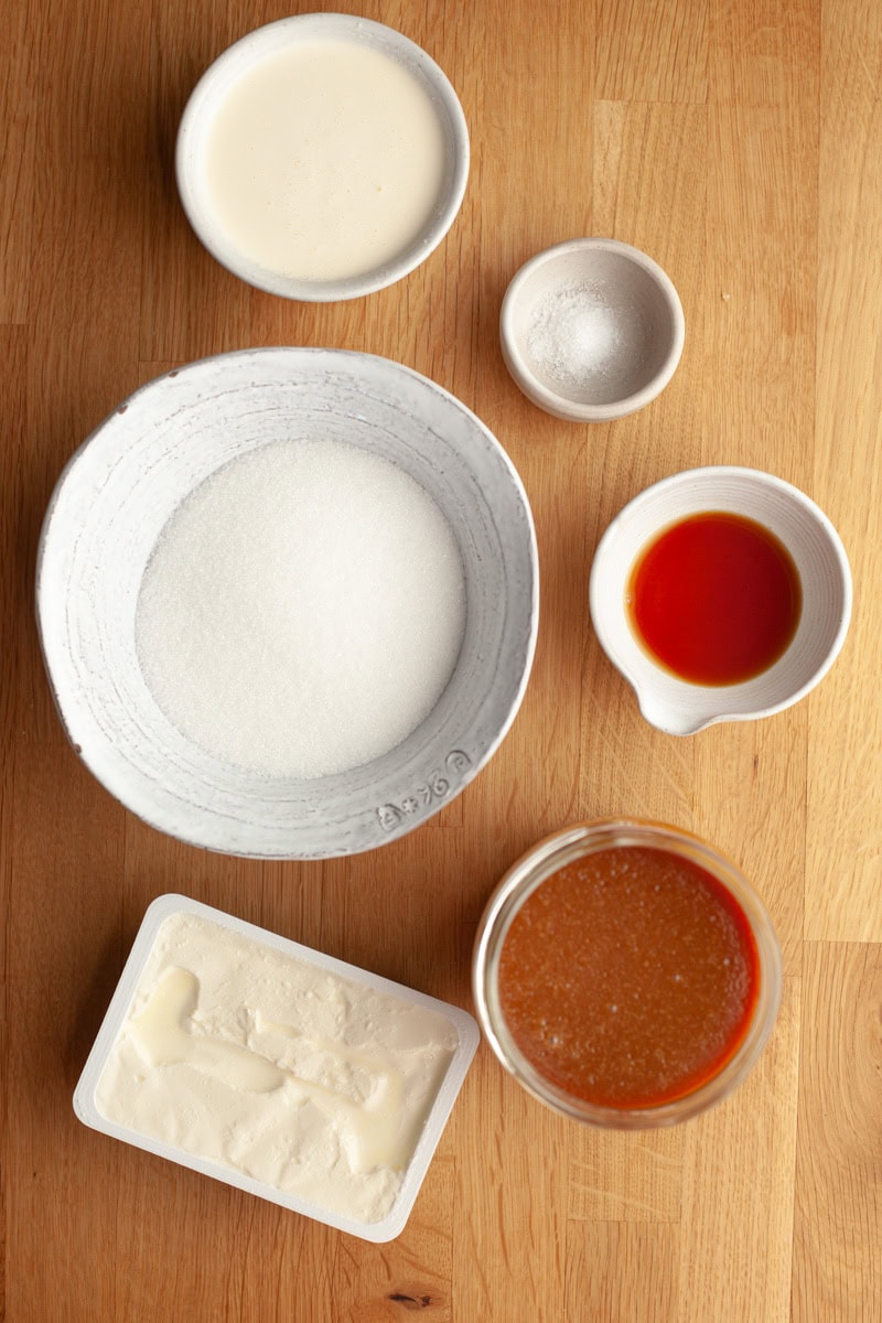 Six white bowls and containers on a wooden surface, holding sugar, cream, salt, vanilla extract, butter, and caramel sauce, arranged neatly for a recipe preparation.