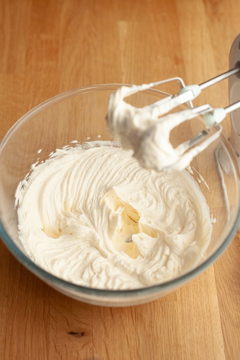 A mixing bowl filled with whipped cream, with a hand mixer coated in the cream resting in the bowl.