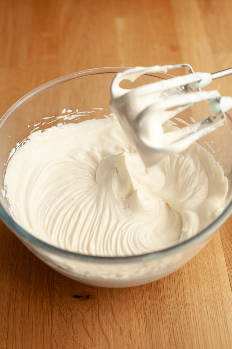 A mixing bowl filled with whipped cream, with a hand mixer coated in the cream resting in the bowl.