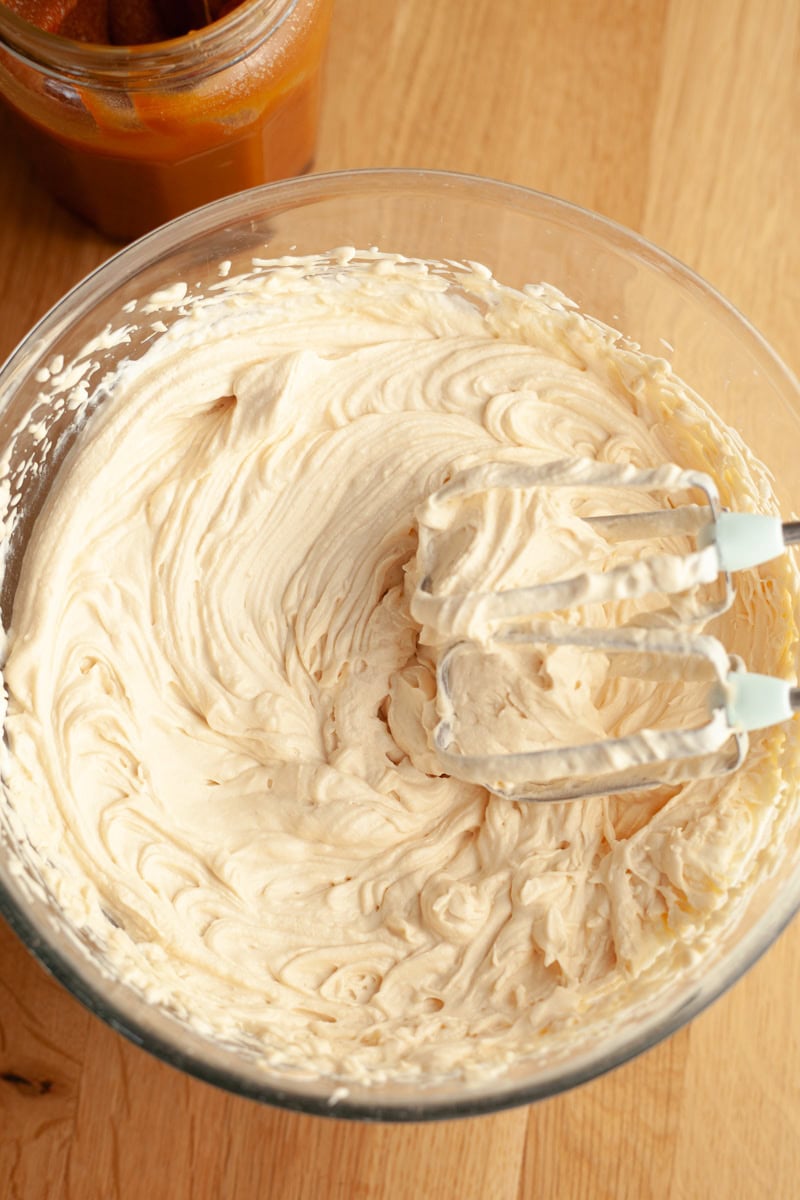 A mixing bowl filled with whipped salted caramel cream cheese frosting, with a hand mixer coated in the frosting resting in the bowl.