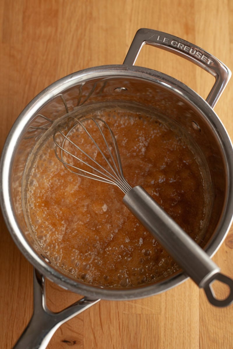 A silver saucepan with a bubbling light brown mixture and a silver whisk.