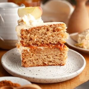 A close-up of a slice of apple layered cake with cream cheese frosting and apple garnish, served on a speckled plate. A fork, spice plate, and more cake are in the background on a wooden table.