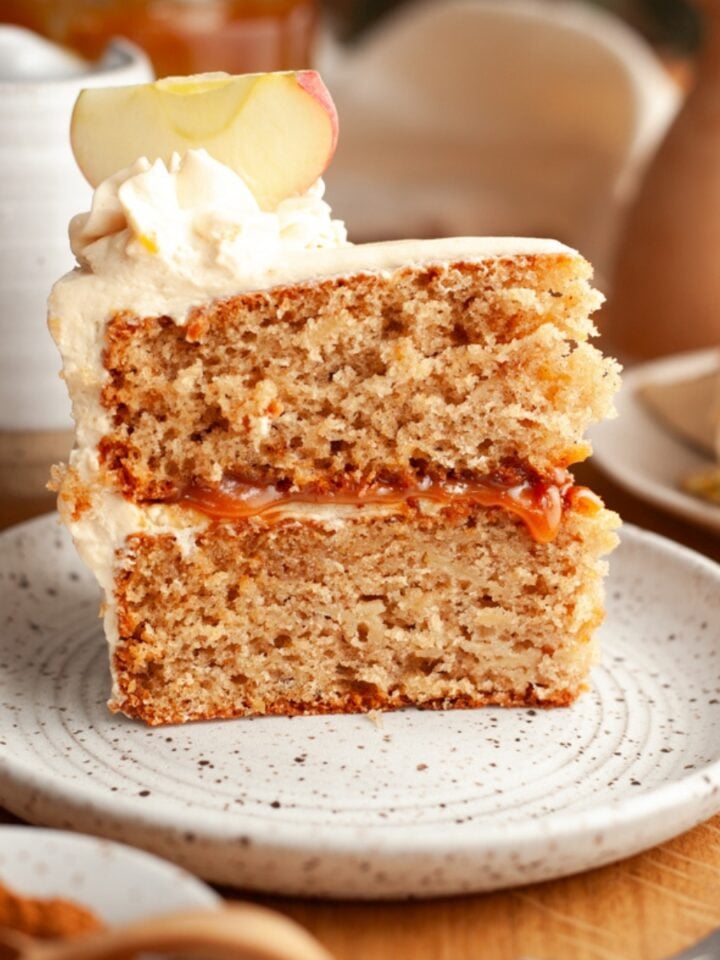 A close-up of a slice of apple layered cake with cream cheese frosting and apple garnish, served on a speckled plate. A fork, spice plate, and more cake are in the background on a wooden table.